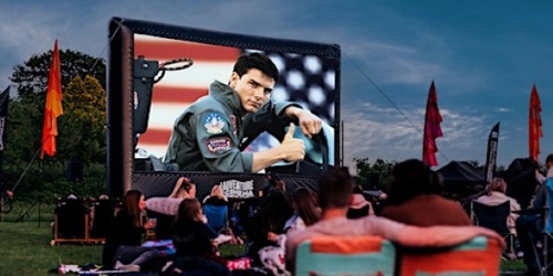 Large cinema screen outdoors showing a scene from Top Gun. People sat in front of it on chairs or blankets watching.