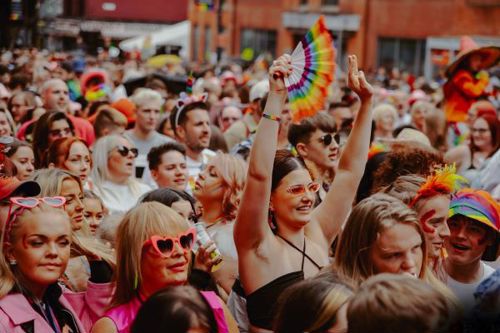 Crowd of people celebrating dressed in bright colours. Girl in the middle has her hands in the air and is holding a rainbow fan.